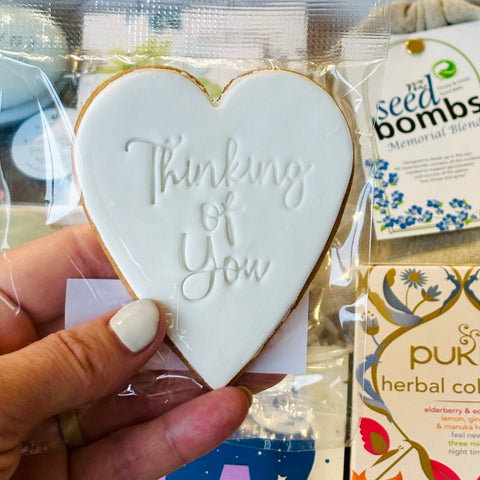 Heart-shaped cookie with 'Thinking of You' text held by a hand, with gift bags and products in the background.