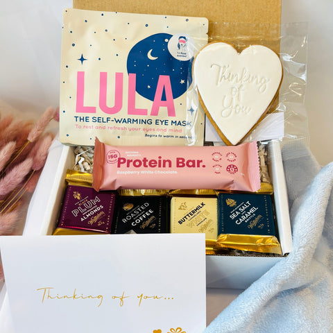 Open gift box with self-warming eye mask, protein bar, and other items on a light background