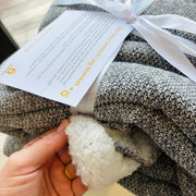 Gray blanket with white fleece inside held by a hand, branded with a visible tag.