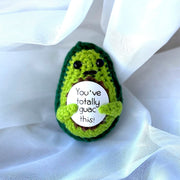 Crocheted avocado-shaped plushie with a humorous message on a white fabric background