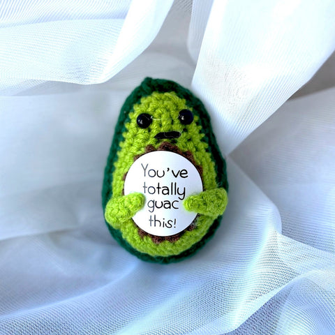 Crocheted avocado-shaped plushie with a humorous message on a white fabric background